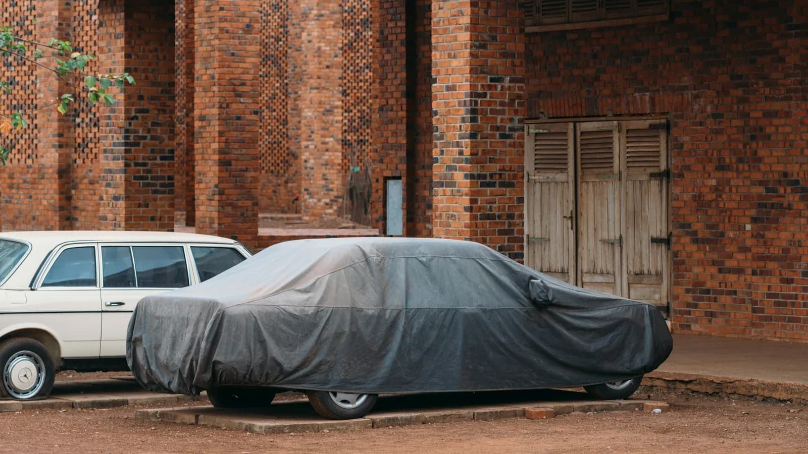 A car under a grey cover parked off the road beside a brick building
