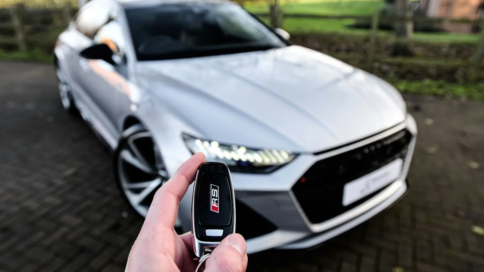 A hand holding a car key in front of a silver car on a driveway